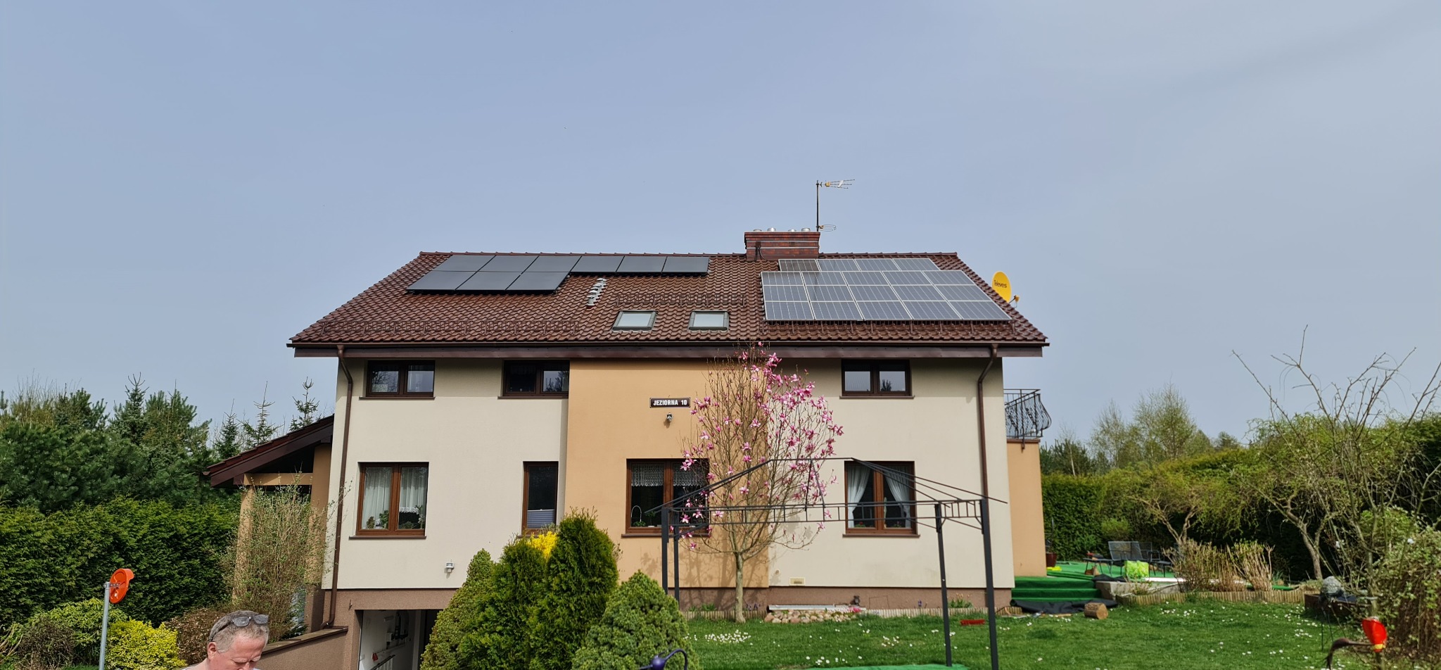 Dom jednorodzinny z brązowym dachem, na którym zamontowano panele słoneczne po obu stronach, z anteną satelitarną i telewizyjną, z tabliczką adresową 'Jeziorna 10' i kwitnącą magnolią na pierwszym...