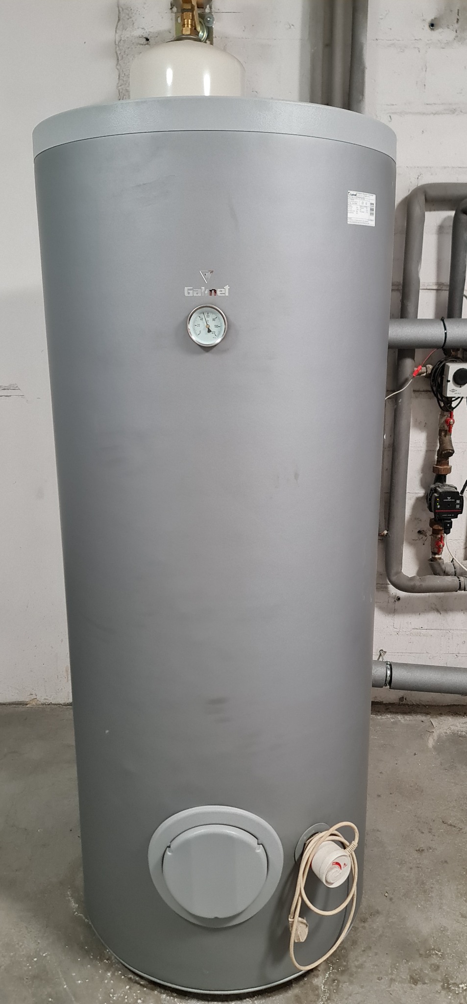 Szary, pionowy zbiornik ciepłej wody użytkowej marki Galmet z termometrem, widoczne rury instalacji grzewczej i przewód zasilający.