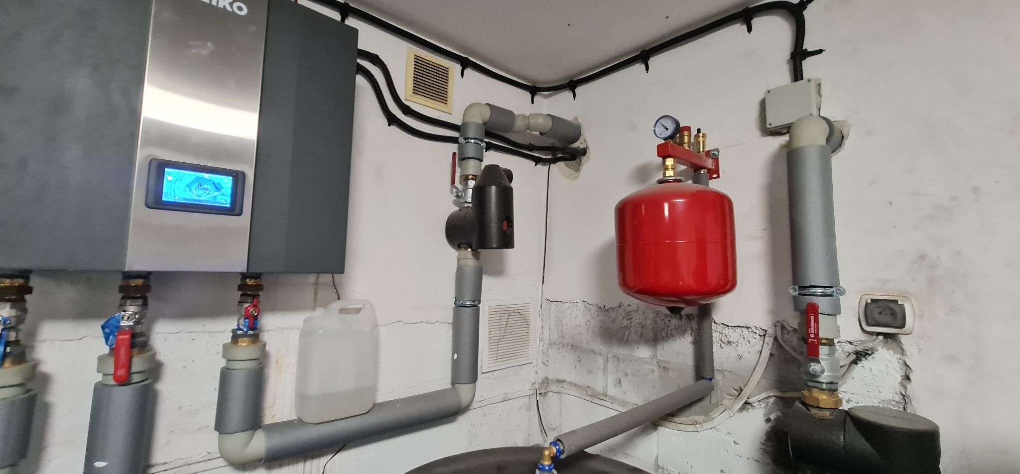 Instalacja pompy ciepła z czerwonym zbiornikiem wyrównawczym i szarymi rurami w pomieszczeniu technicznym w Rumi.