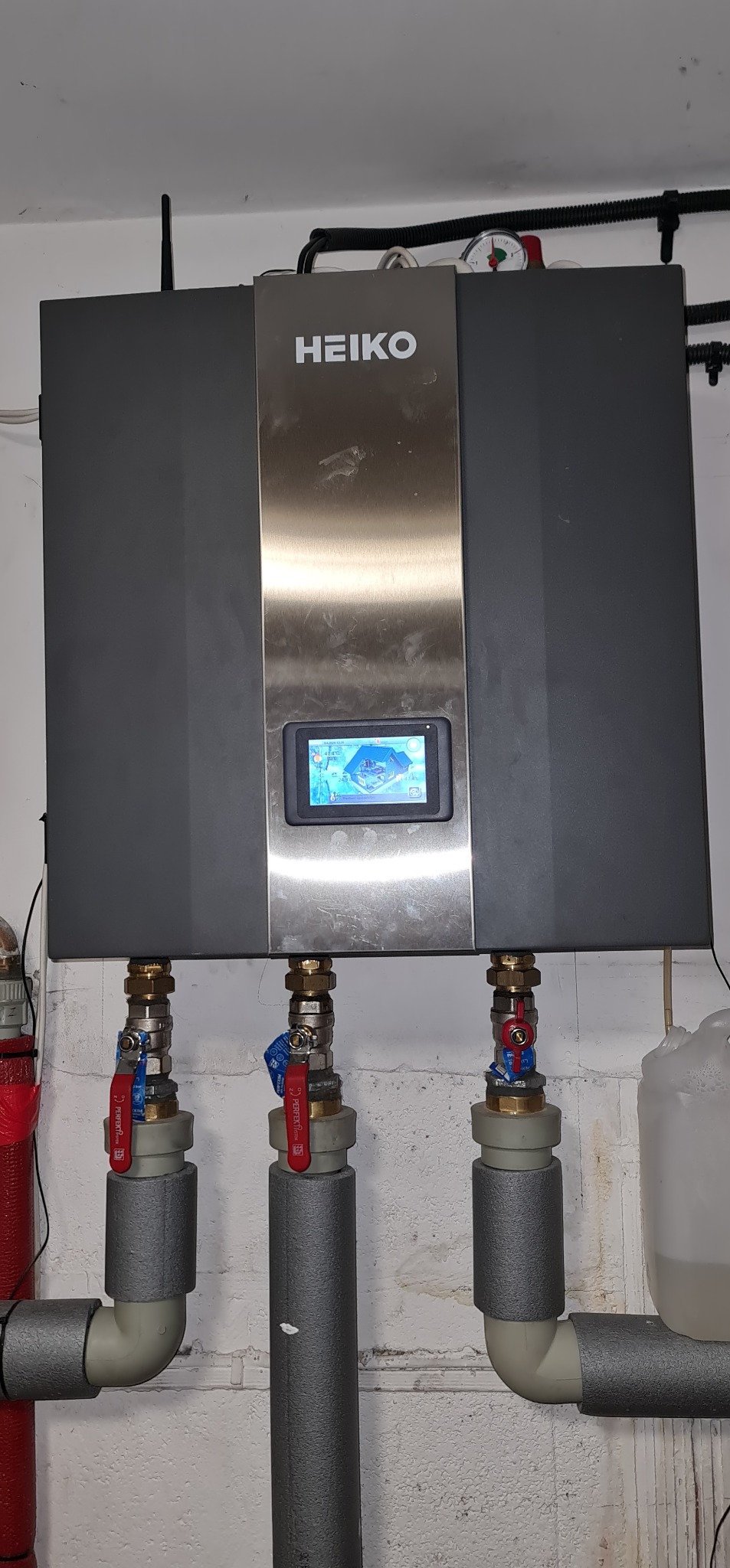 Szary panel sterowania HEIKO z ekranem dotykowym wyświetlającym schemat domu i instalacji, widoczne zawory i izolowane rury instalacyjne na tle białej ściany.