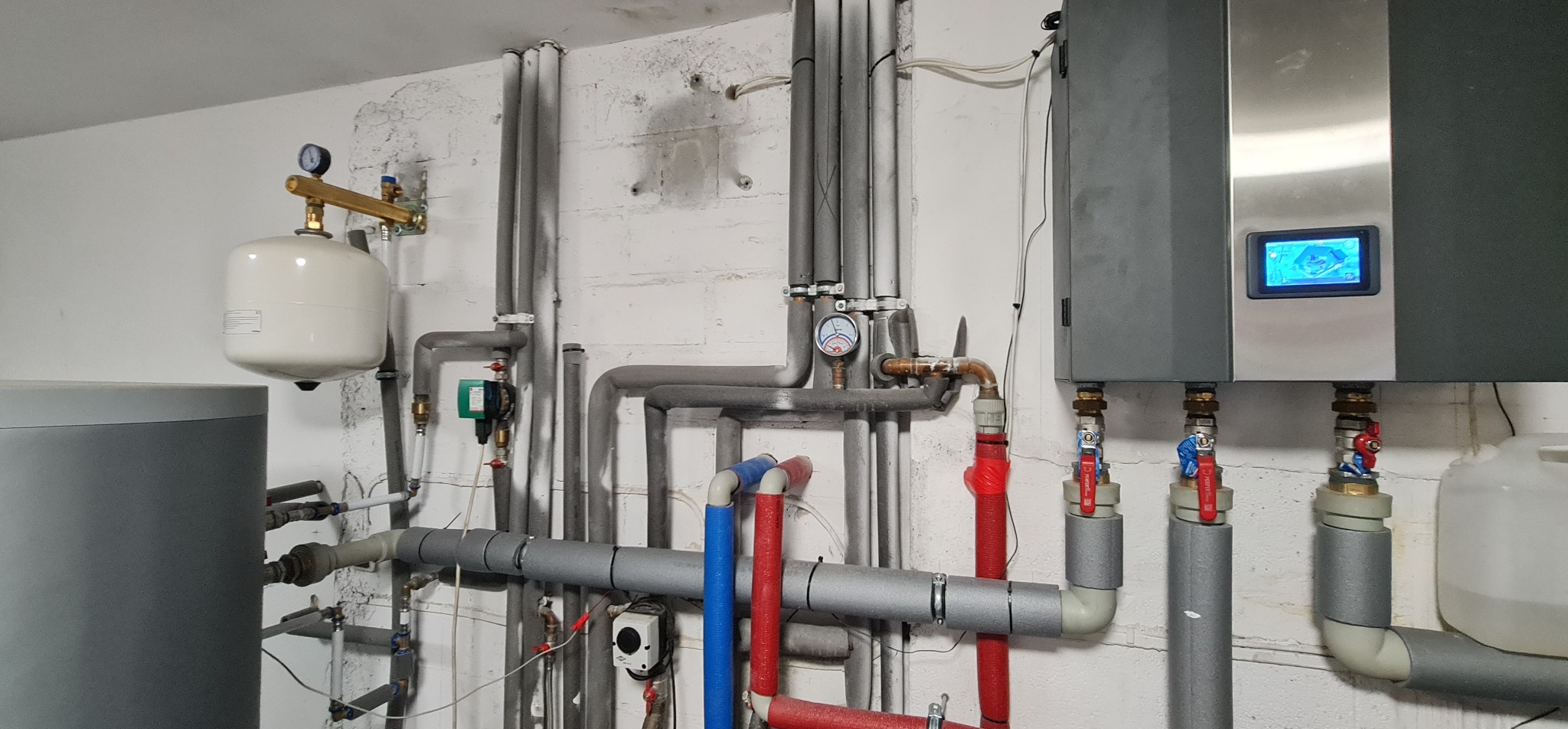 Węzeł cieplny z szarym zbiornikiem buforowym, białym naczyniem przeponowym, licznymi rurami z izolacją termiczną, zaworami i elektronicznym sterownikiem z wyświetlaczem na białej, lekko zabrudzonej...