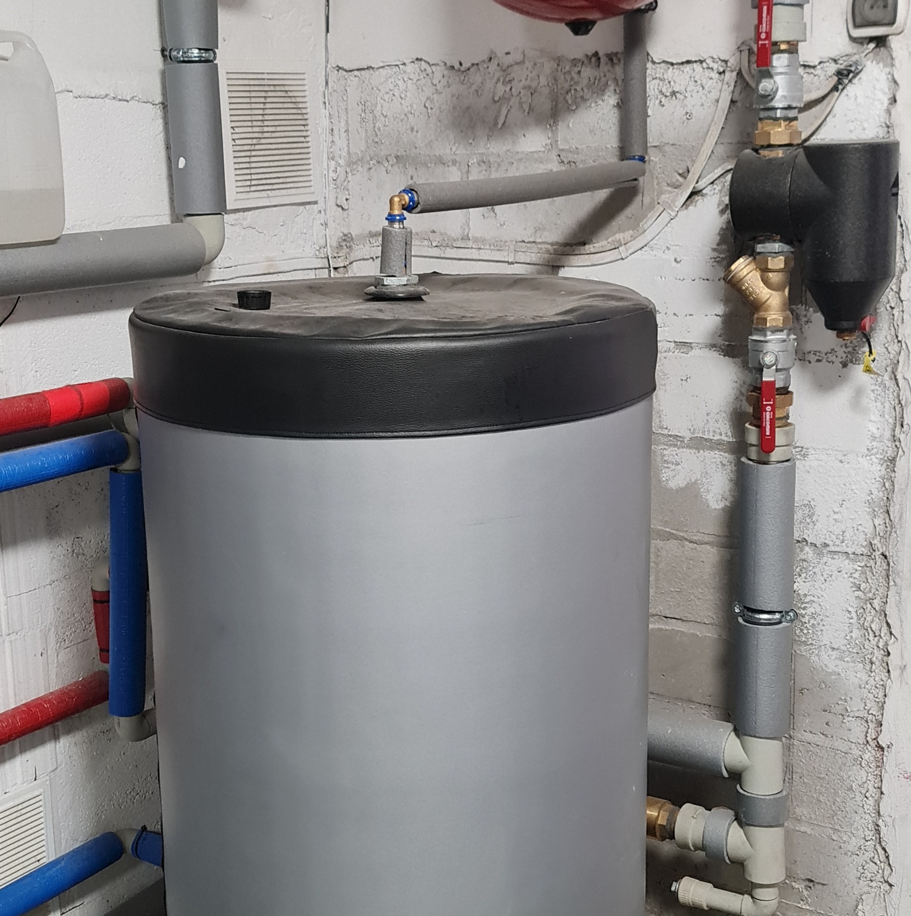 Szary bojler z czarną pokrywą, podłączony do instalacji z rurami w kolorze czerwonym i niebieskim, z widocznymi zaworami i izolacją termiczną na tle ściany z białej cegły.