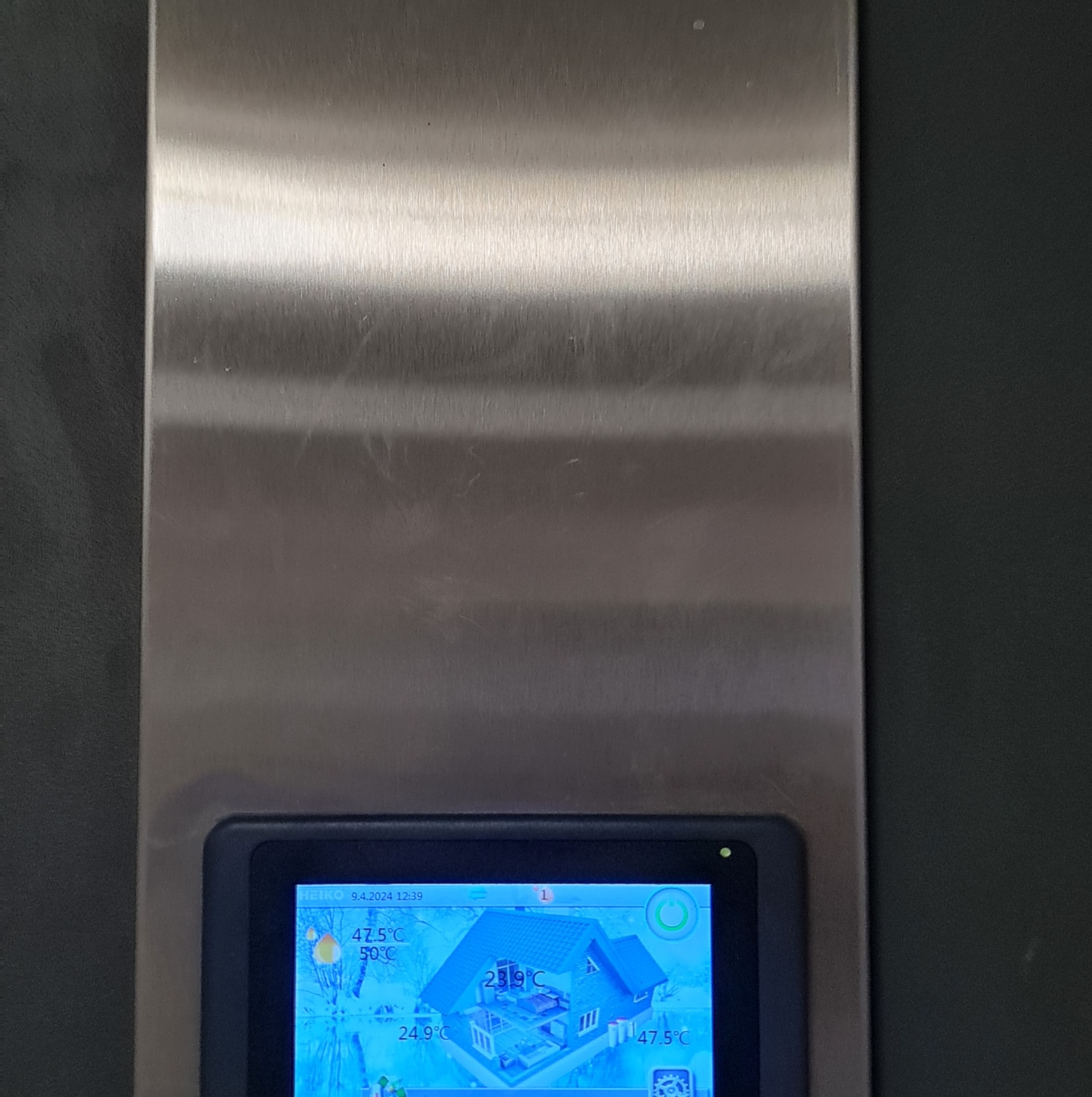 Ekran dotykowy panelu sterowania z interfejsem graficznym przedstawiającym schemat domu i odczyty temperatur, wbudowany w metalową obudowę.