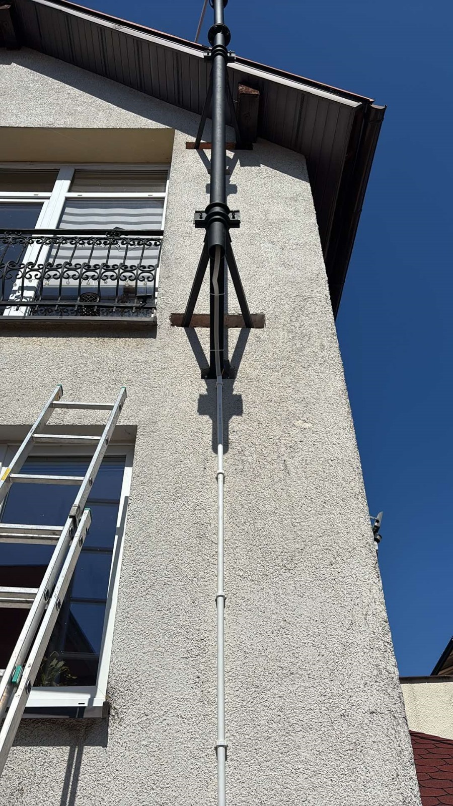 Montaż instalacji odgromowej na elewacji domu z oknem i balkonem. Widoczna drabina oparta o ścianę. Błękitne niebo w tle.