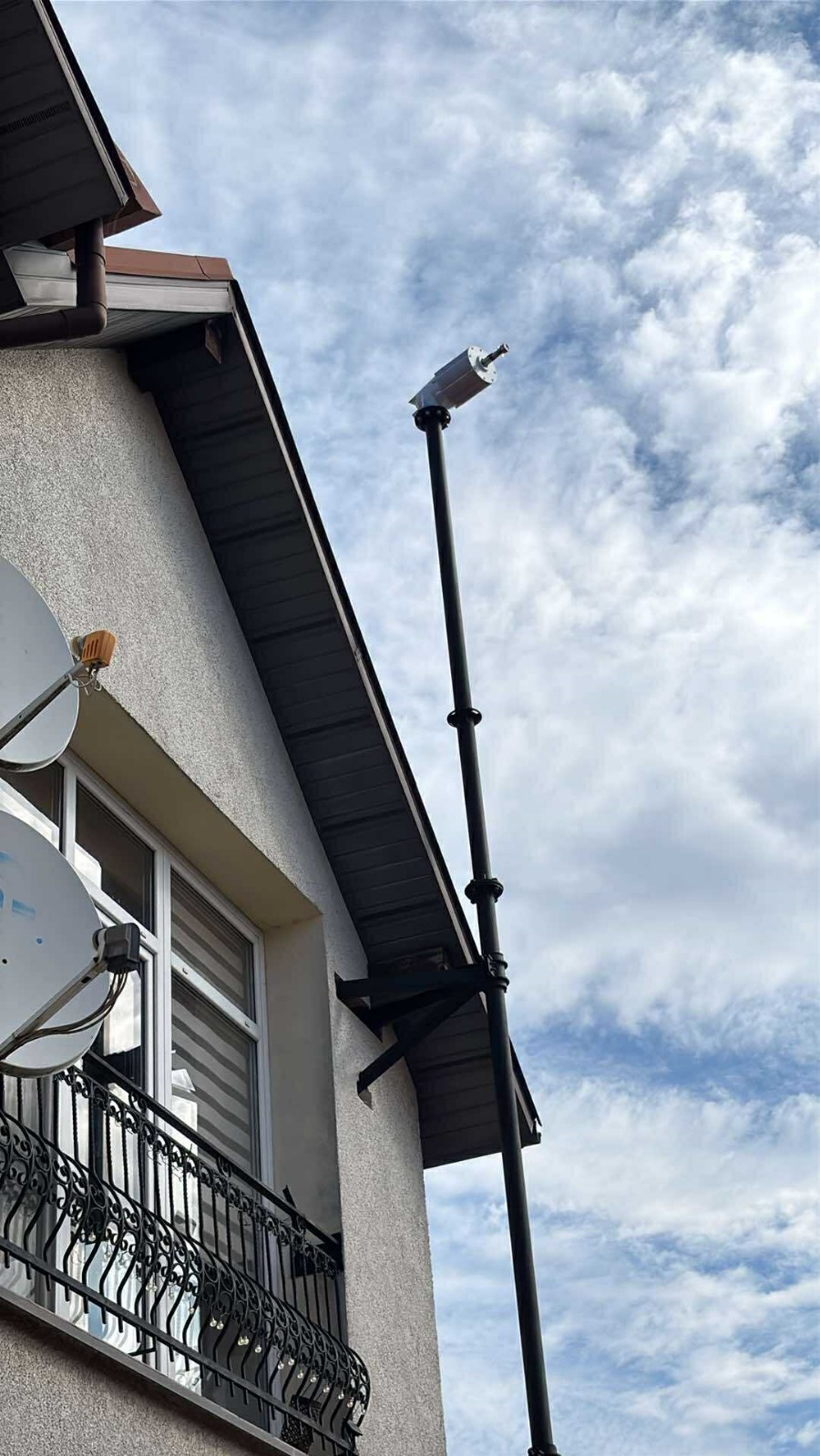 Montaż małej turbiny wiatrowej na elewacji domu z balkonem i anteną satelitarną, na tle błękitnego nieba z chmurami.