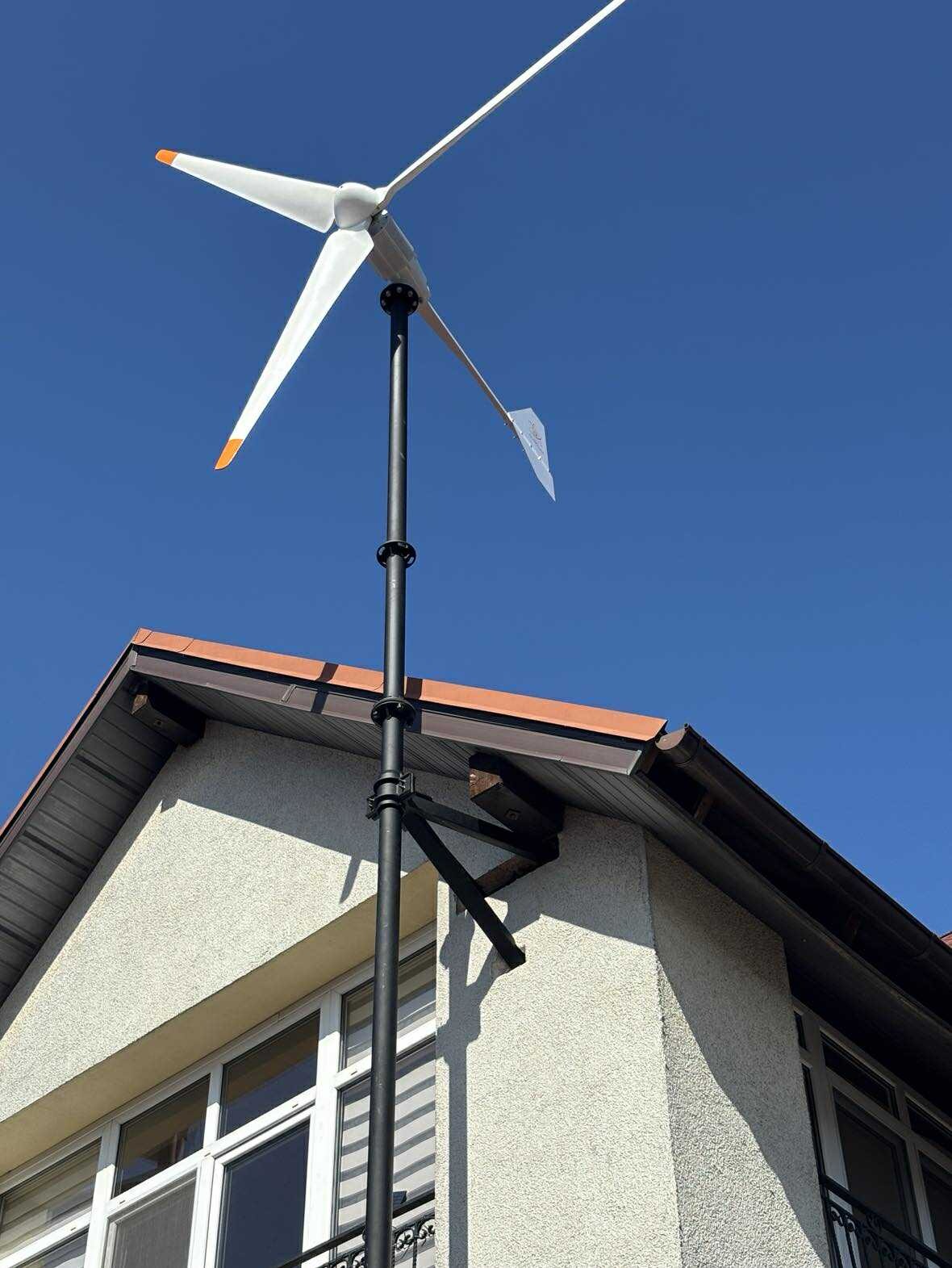 Turbina wiatrowa zamontowana na budynku mieszkalnym, widoczna na tle błękitnego nieba. Trzy łopaty z pomarańczowymi końcówkami. Widoczne okna budynku.