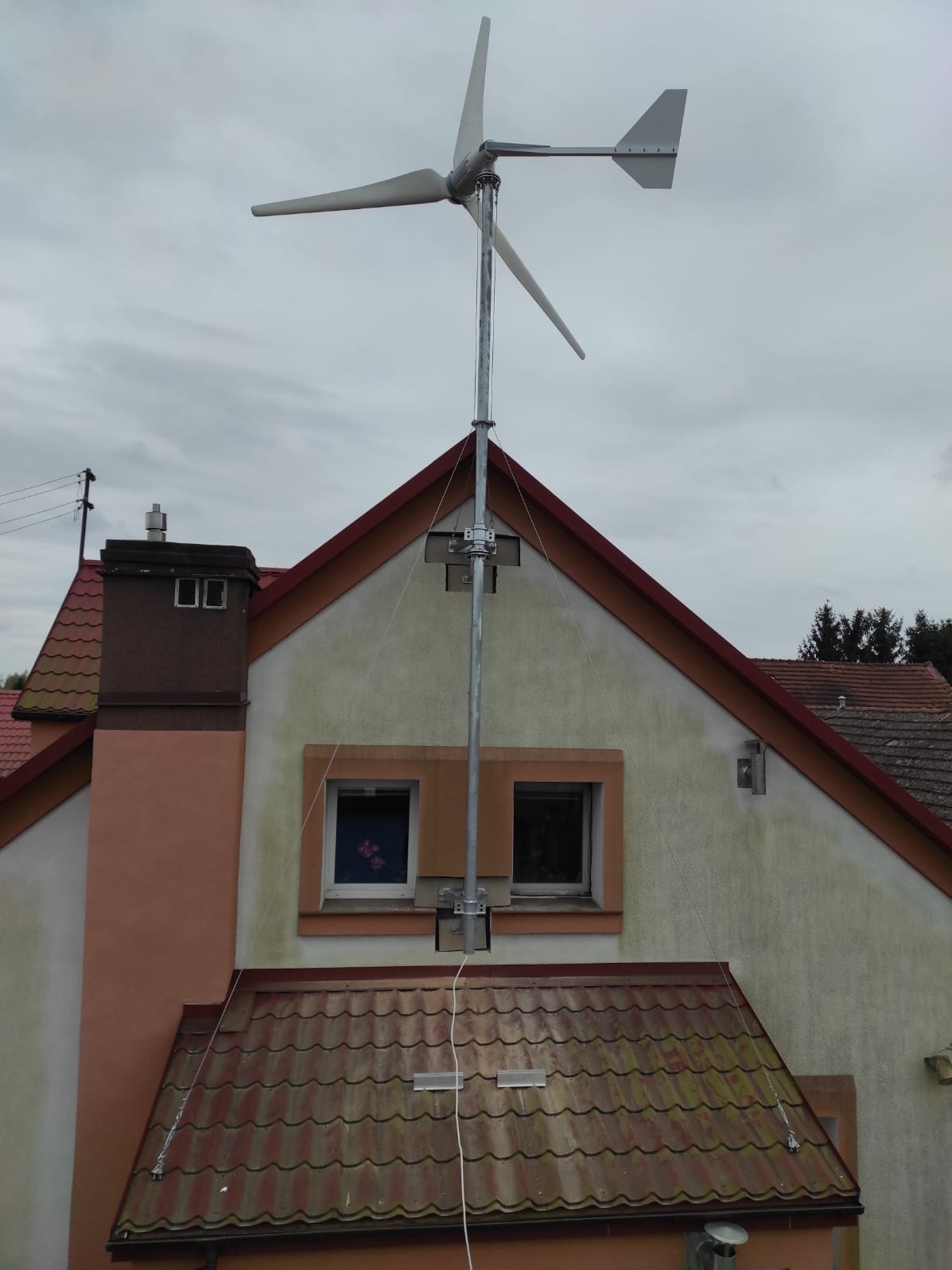 Turbina wiatrowa zamontowana na dachu dwupiętrowego domu z kominem. Metalowa konstrukcja z białymi śmigłami na tle pochmurnego nieba.