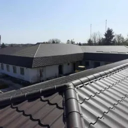 Widok z góry na budynek z ciemnoszarą blachodachówką, z widocznym fragmentem brązowej blachodachówki na pierwszym planie i żółtym samochodem dostawczym w tle.