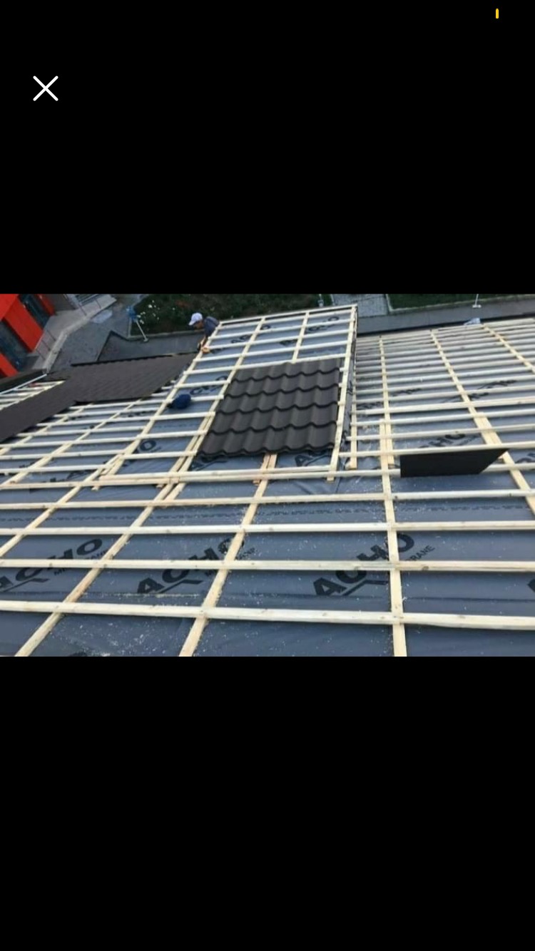 Montaż metalowej dachówki na drewnianej konstrukcji dachu pokrytej membraną, widoczny pracownik w oddali.