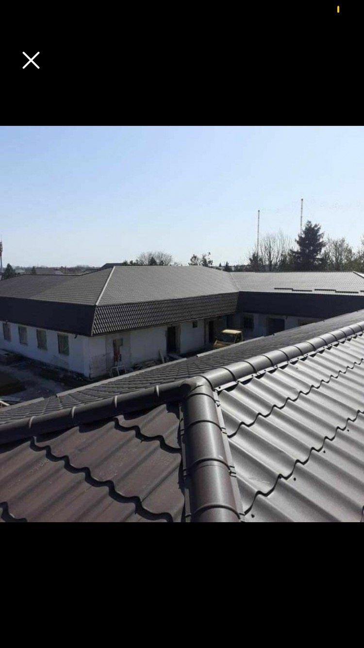 Widok z góry na budynek z ciemnoszarą blachodachówką, z widocznym fragmentem brązowej blachodachówki na pierwszym planie i żółtym samochodem dostawczym w tle.