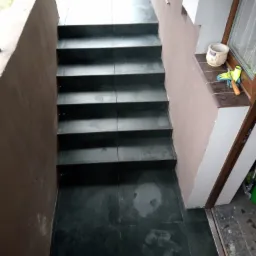 Schody zewnętrzne wyłożone ciemną płytką, prowadzące do wejścia, z widocznymi śladami po pracy, obok okna leżą rękawice robocze i śrubokręt.