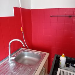 MB-complex - Kuchnia po remoncie: Ściany wyłożone czerwonymi płytkami, biała góra, stalowy zlew z baterią, widoczny fragment szafki pod zlewem oraz akcesoria do czyszczenia.