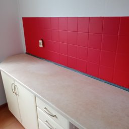 MB-complex - Kuchnia po remoncie: czerwone płytki nad blatem, jasne szafki i widoczne gniazdko elektryczne.