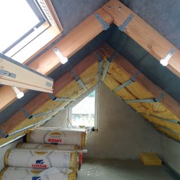 MB-complex - Poddasze w trakcie ocieplania: widoczne krokwie z metalowymi uchwytami, żółta wełna mineralna pomiędzy nimi, folia paroizolacyjna na suficie oraz rolki wełny Ursa Glasswool Gold 35 na podłodze.