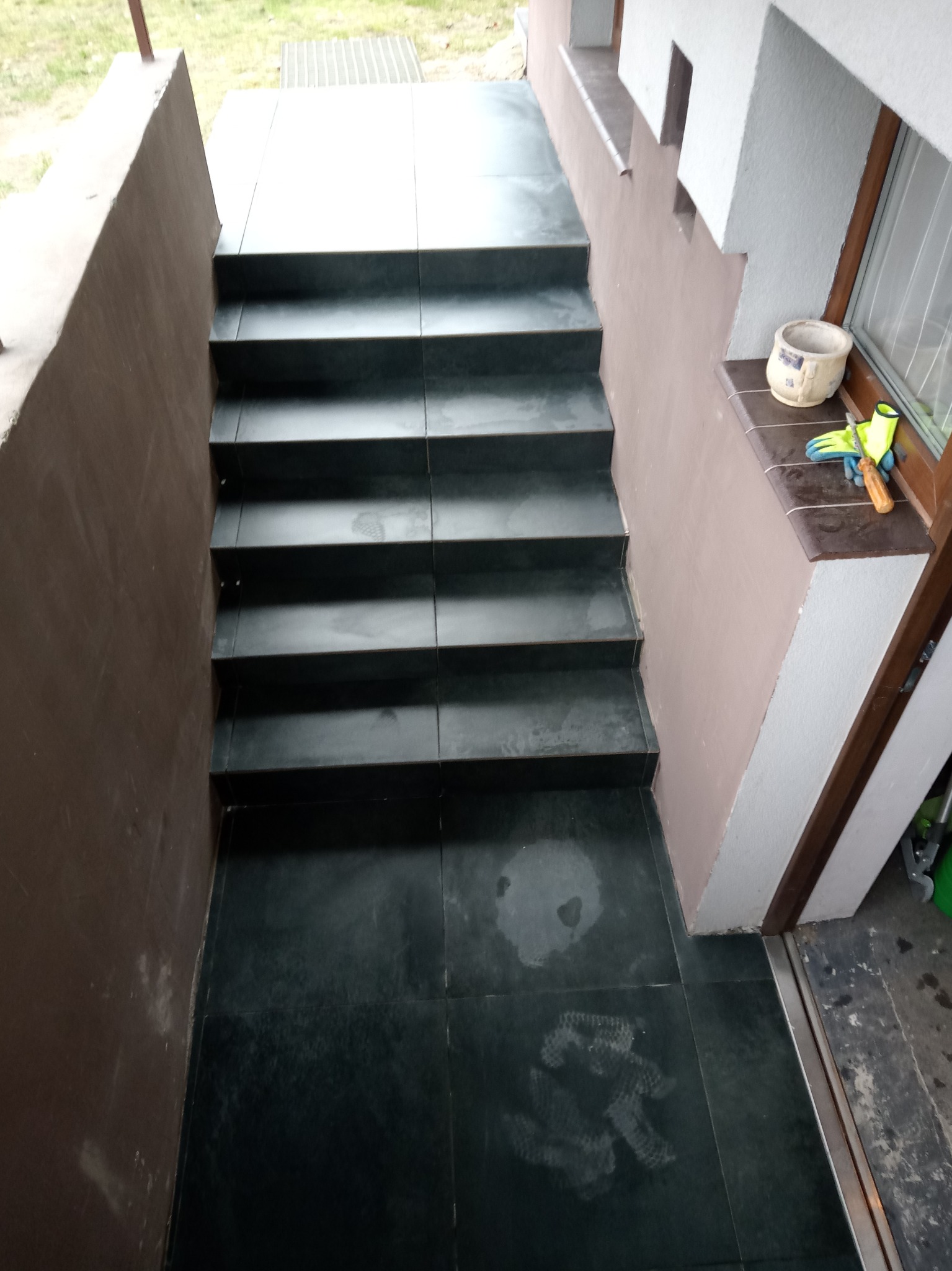 Schody zewnętrzne wyłożone ciemną płytką, prowadzące do wejścia, z widocznymi śladami po pracy, obok okna leżą rękawice robocze i śrubokręt.