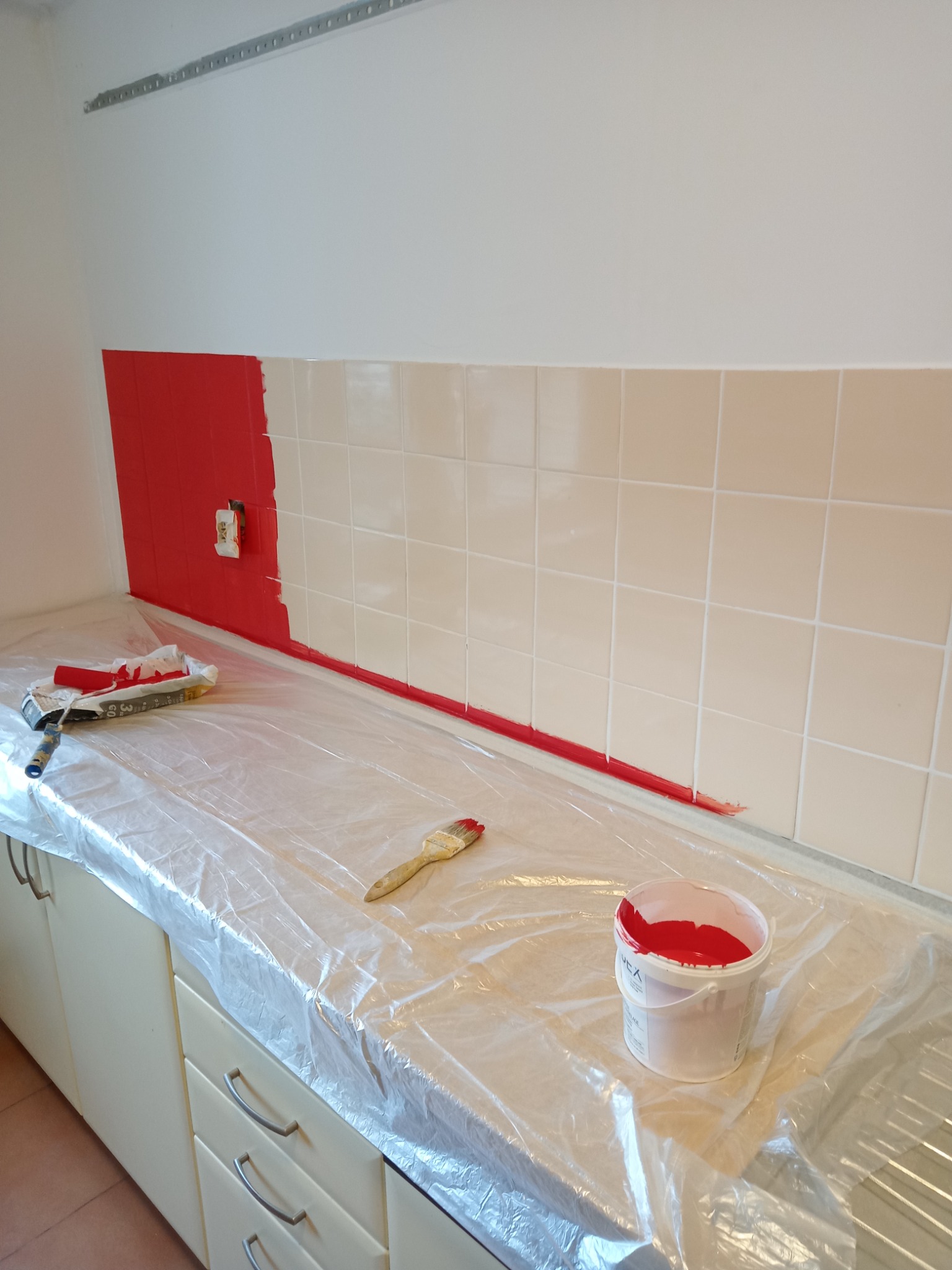 Częściowo pomalowane, czerwone płytki nad blatem w kuchni w Jeżowem, zabezpieczonym folią, widoczny wałek i pędzel malarski oraz wiaderko z farbą.