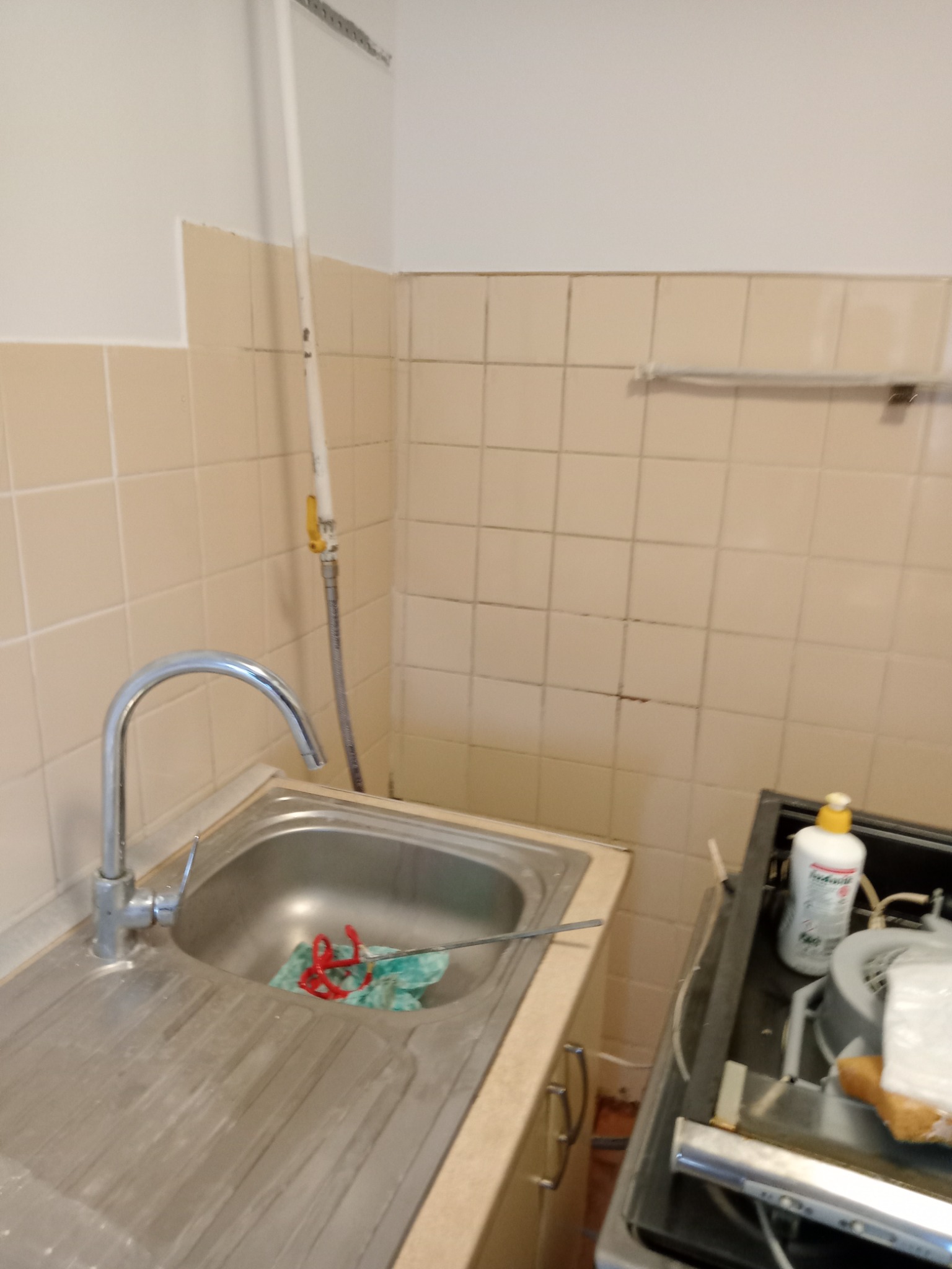 Kuchnia przed remontem: widok na zlew ze śladami po remoncie, fragment kuchenki i ściany z beżowymi kaflami, widoczna rura gazowa.