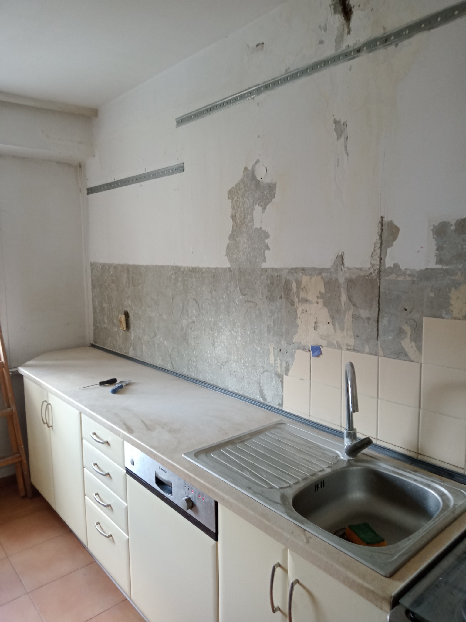 Kuchnia w trakcie remontu: ściana z usuniętą częścią starej okładziny, widoczne metalowe listwy montażowe, blat z narzędziami i zlewem, w tle drabina.