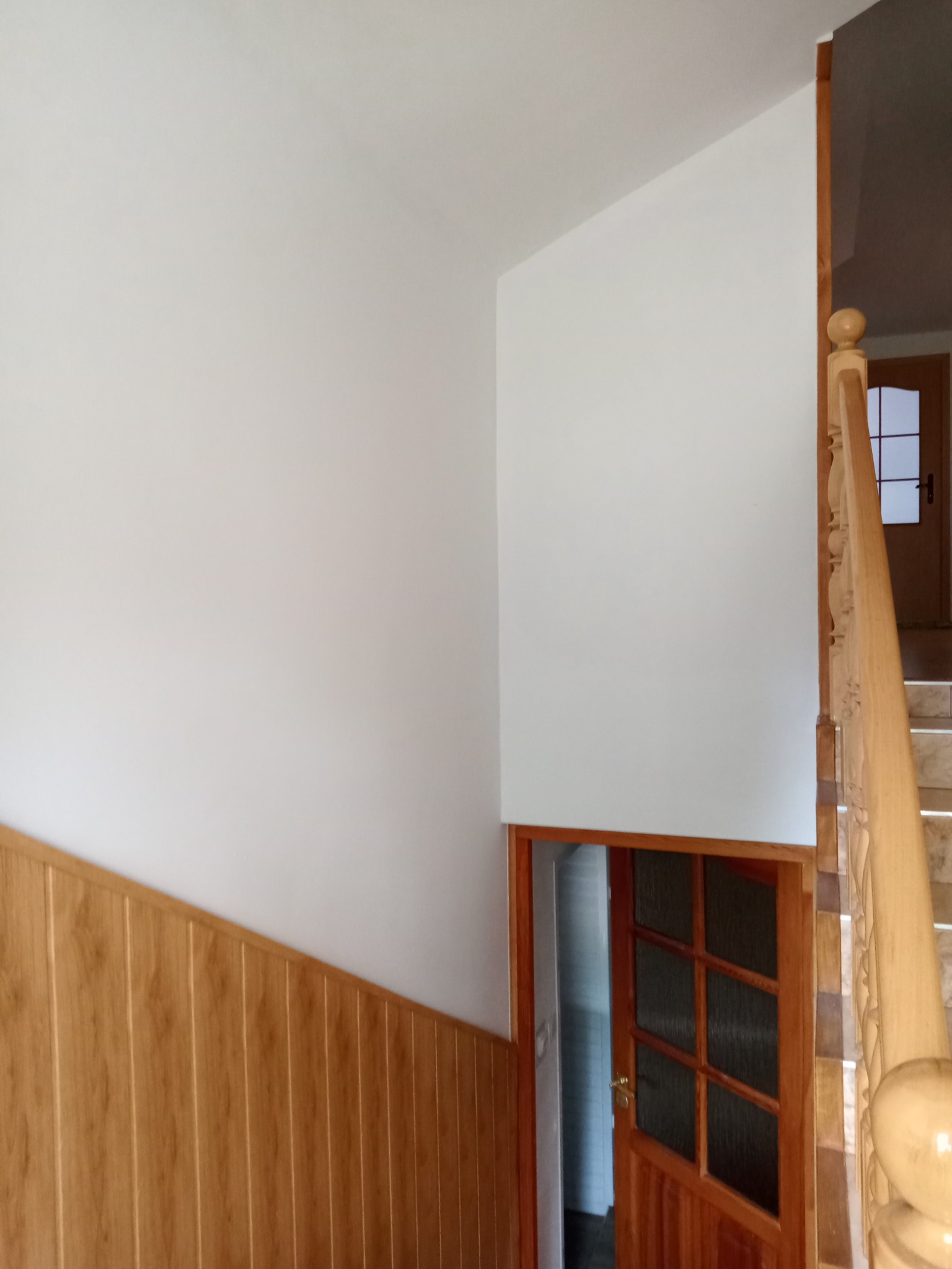 Świeżo pomalowana, biała ściana w korytarzu z drewnianą boazerią, drewnianymi schodami i drzwiami z szybkami. Widoczne detale wykończenia schodów i drzwi.