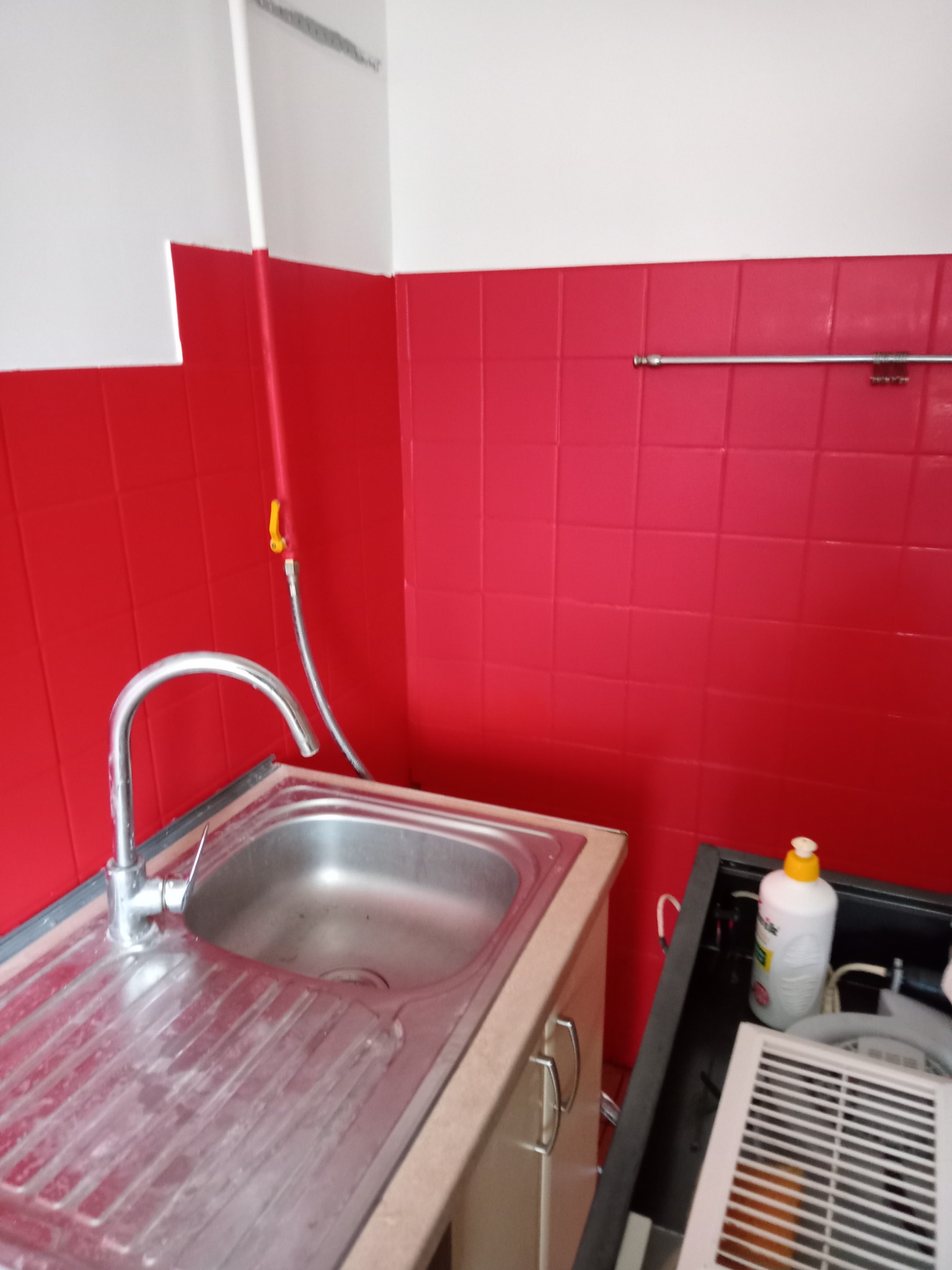 Kuchnia po remoncie: Ściany wyłożone czerwonymi płytkami, biała góra, stalowy zlew z baterią, widoczny fragment szafki pod zlewem oraz akcesoria do czyszczenia.