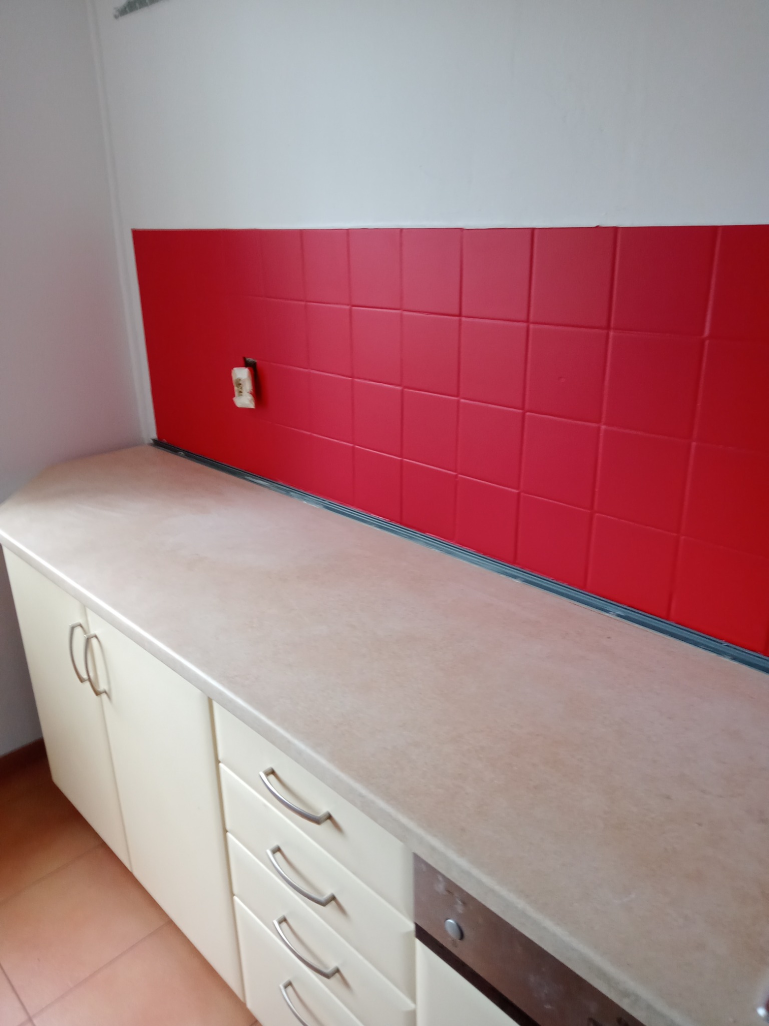 Kuchnia po remoncie: czerwone płytki nad blatem, jasne szafki i widoczne gniazdko elektryczne.