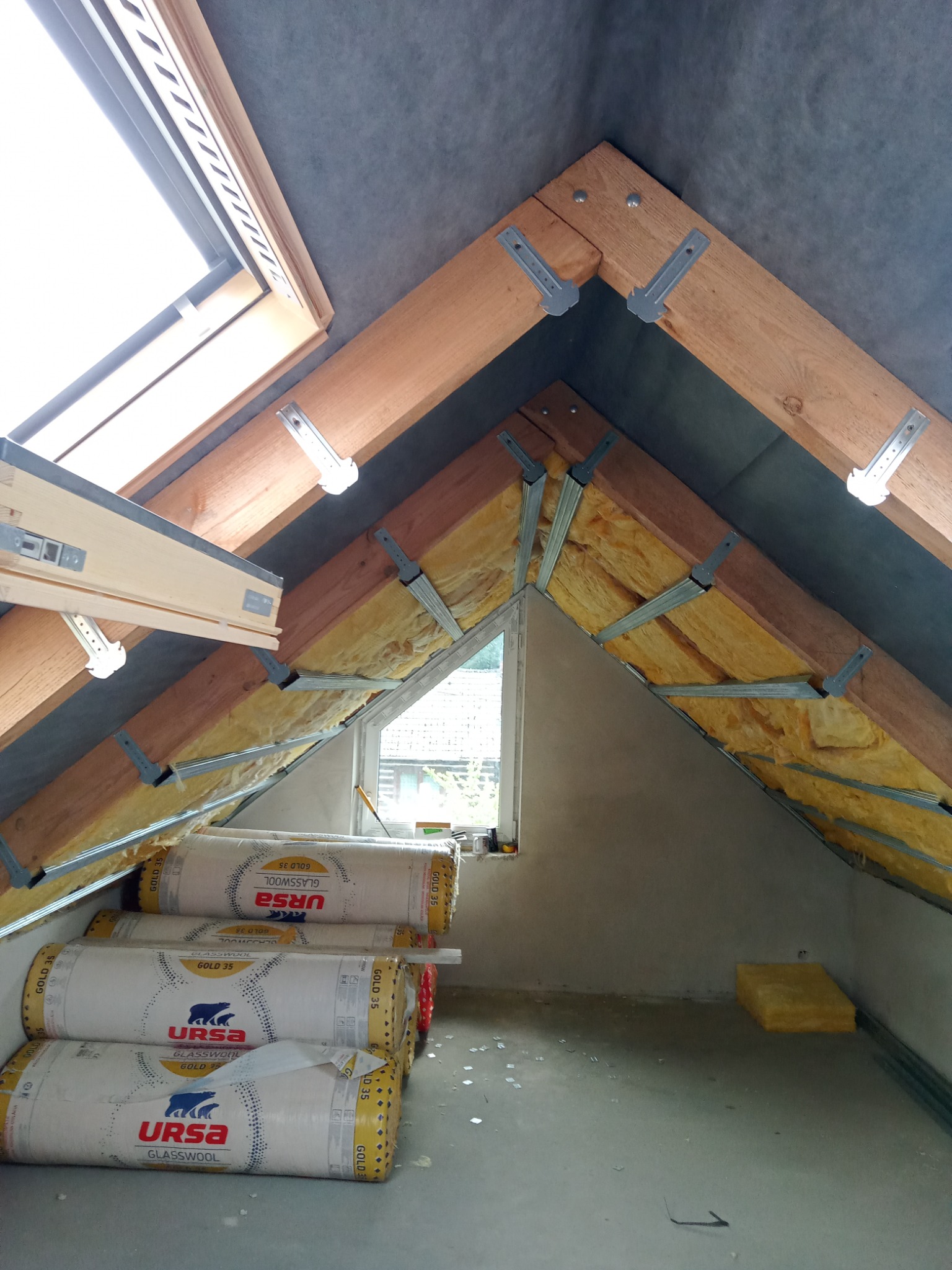 Poddasze w trakcie ocieplania: widoczne krokwie z metalowymi uchwytami, żółta wełna mineralna pomiędzy nimi, folia paroizolacyjna na suficie oraz rolki wełny Ursa Glasswool Gold 35 na podłodze.