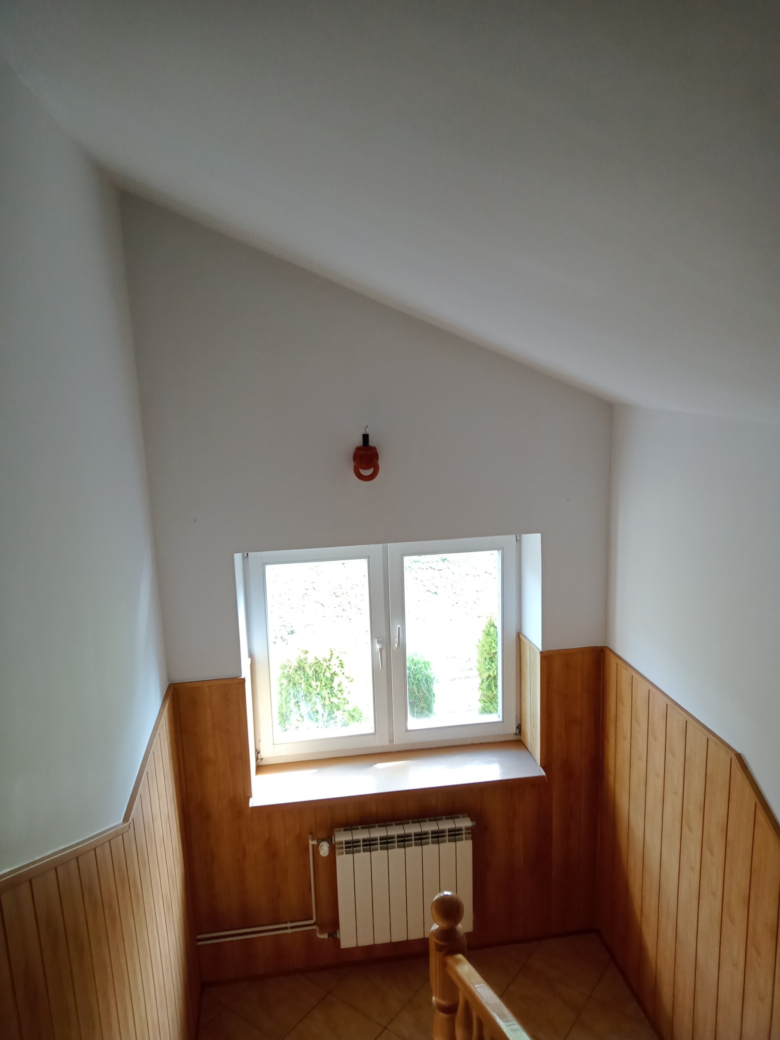 Poddasze z oknem, drewnianą boazerią, grzejnikiem i schodami, widok na zieleń za oknem.