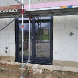 Ciemne okno zamontowane w surowej ścianie z bloczk&oacute;w, widoczne rusztowanie i fragment izolacji, teren wok&oacute;ł w trakcie prac budowlanych.