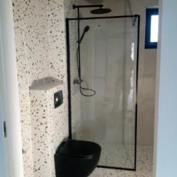 Nowoczesna łazienka z czarną armaturą: toaleta podwieszana, kabina prysznicowa z czarną ramą i deszczownicą, płytki lastryko na ścianach i podłodze, wbudowana półka ścienna.