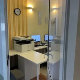 Jasne biuro rachunkowe z dwoma stanowiskami komputerowymi, drukarką na białym biurku i zegarem na ścianie z tapetą w pionowe pasy, widok zza otwartych drzwi.