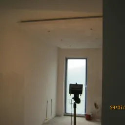 Wnętrze w trakcie prac przygotowawczych do malowania, ściany po szpachlowaniu, widoczna lampa halogenowa oświetlająca pomieszczenie, okno zabezpieczone folią.