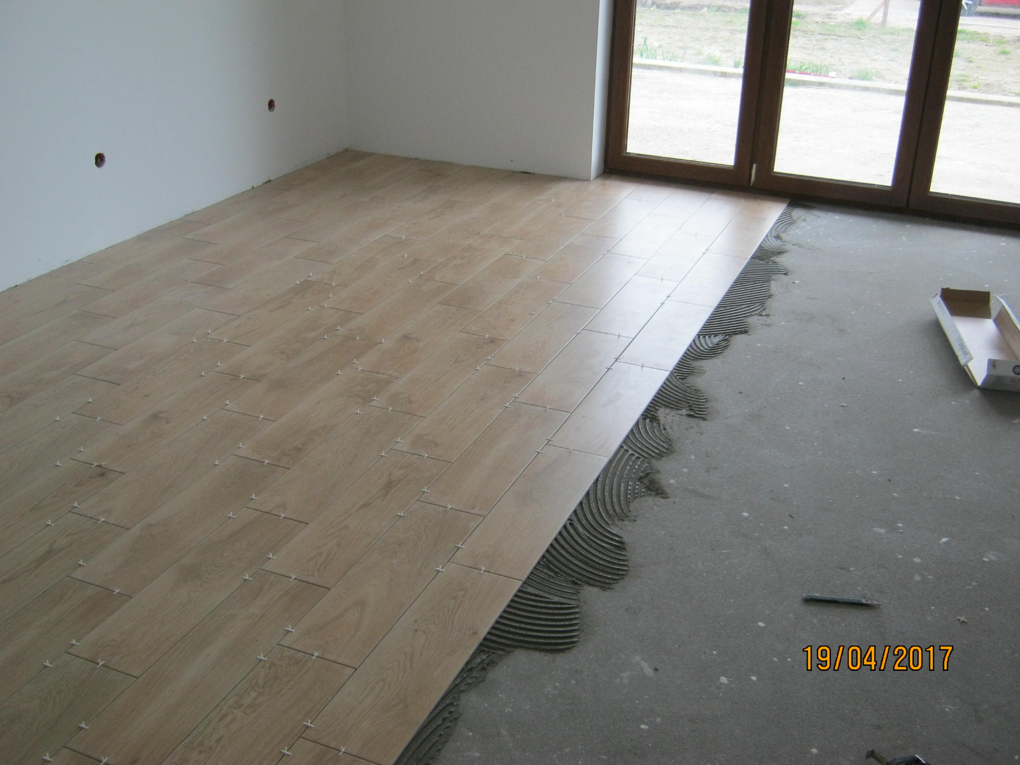 Układanie podłogi z płytek imitujących drewno, widoczna warstwa kleju i krzyżyki dystansowe, pomieszczenie w stanie surowym z oknem.