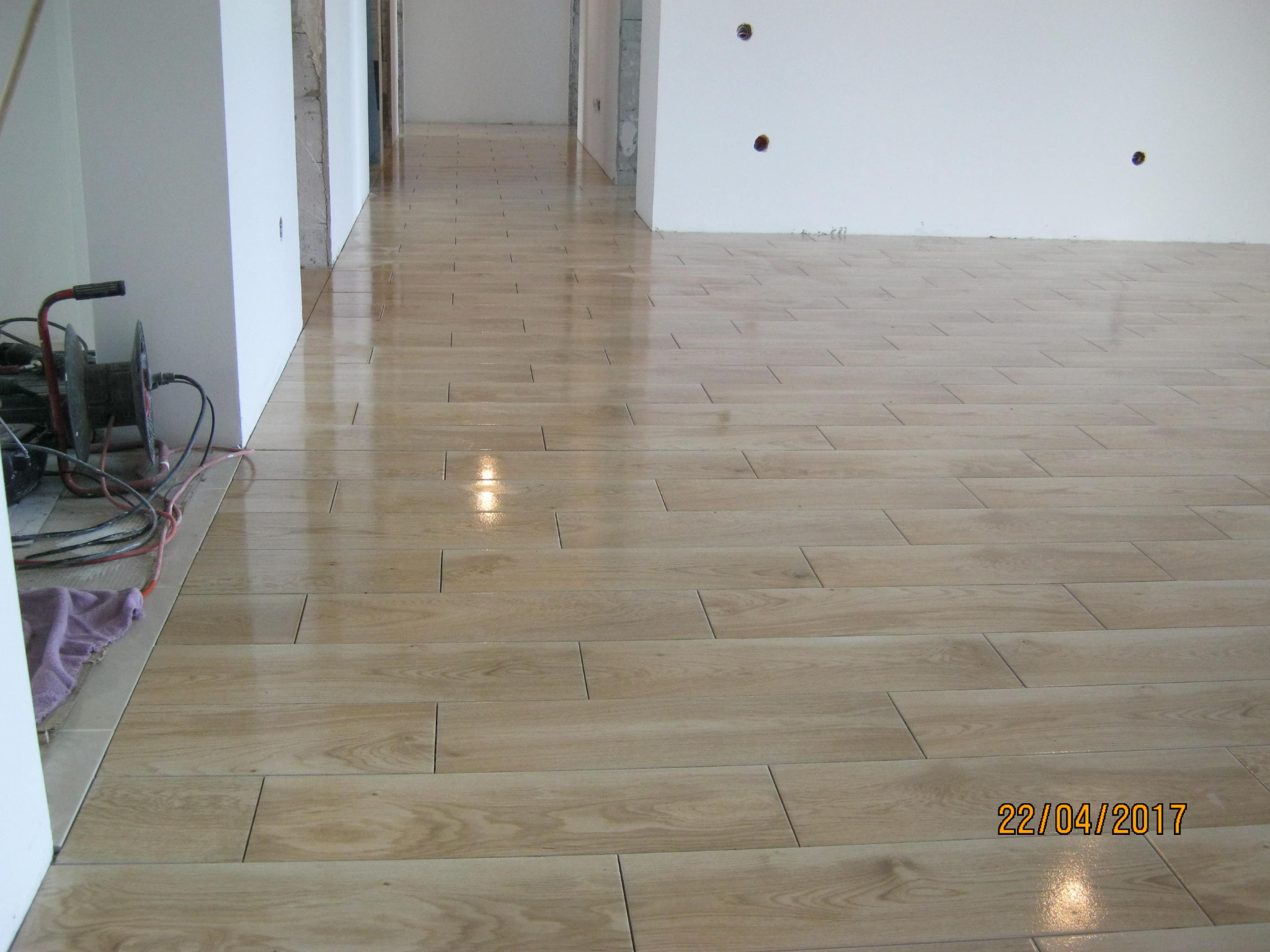 Świeżo położona, błyszcząca podłoga z płytek imitujących drewno w długie deski, widoczne narzędzia pracy i kabel zasilający przy ścianie.