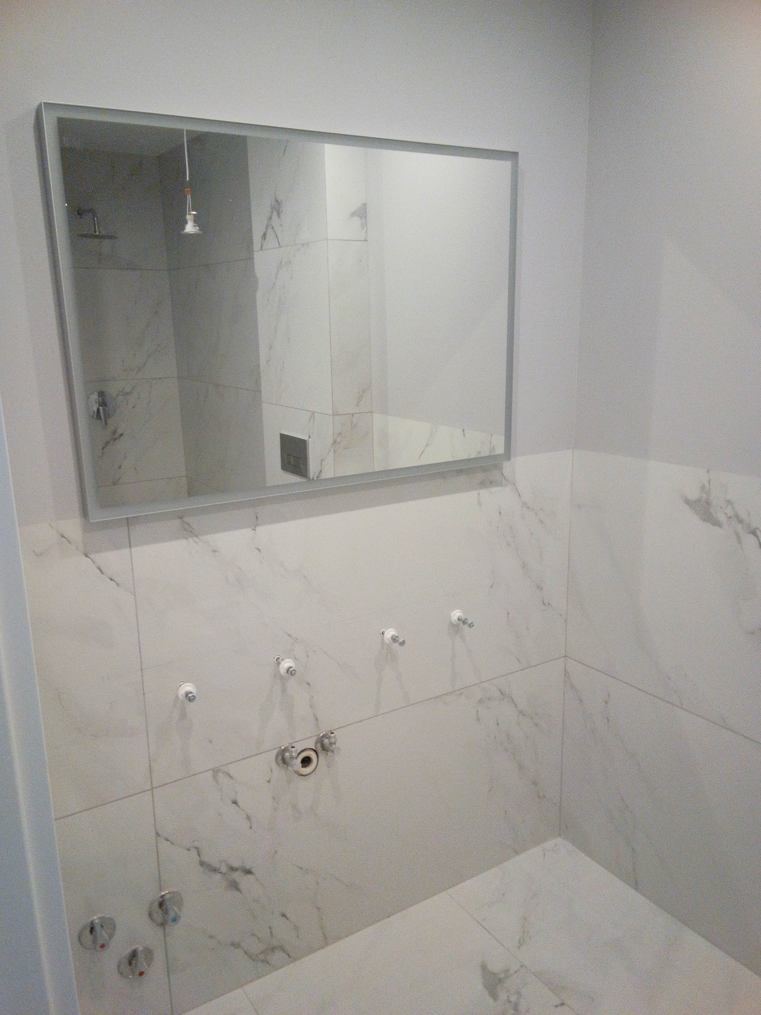 Białe płytki imitujące marmur na ścianach łazienki, widoczne wystające rury i przyłącza, lustro z odbiciem kabiny prysznicowej.