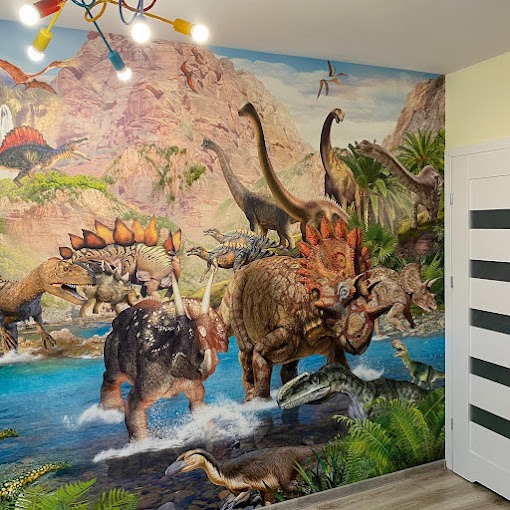 Fototapeta w pokoju dziecięcym z motywem dinozaurów na tle górskiego krajobrazu, oświetlona kolorową lampą sufitową.