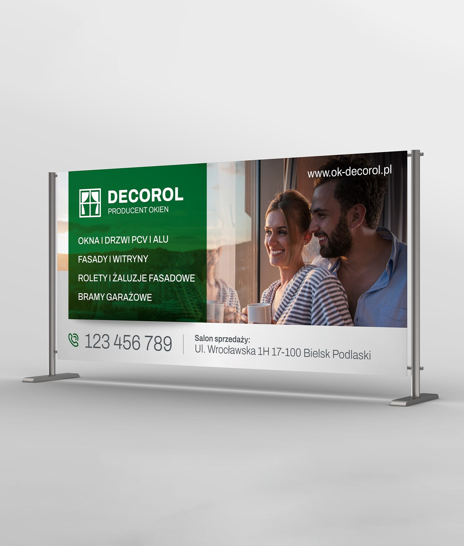 Baner reklamowy firmy Decorol, producenta okien, prezentujący ofertę okien, drzwi, fasad, rolet i bram garażowych, z uśmiechniętą parą w tle i adresem salonu sprzedaży.