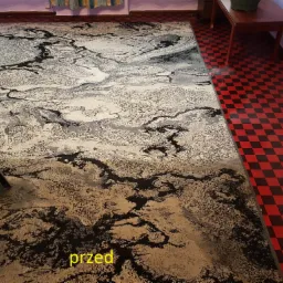 Dywan z abstrakcyjnym wzorem, widoczna różnica w czystości między częścią wyczyszczoną a zabrudzoną, urządzenie do czyszczenia dywanów w lewym dolnym rogu, podłoga w czerwoną szachownicę w tle.