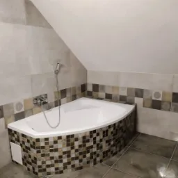 Narożna wanna obudowana mozaiką w odcieniach brązu i beżu, z chromowaną baterią i słuchawką prysznicową na tle jasnych, beżowych płytek w łazience na poddaszu.