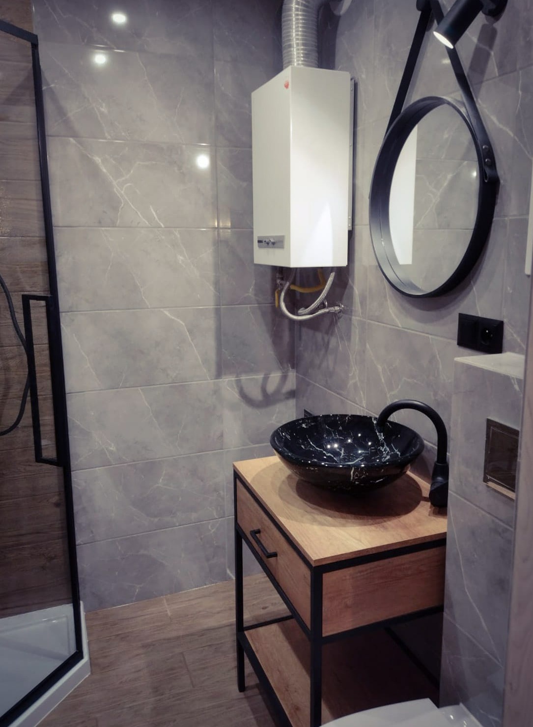 Nowoczesna łazienka z szarymi płytkami imitującymi marmur, czarną umywalką nablatową na drewnianej szafce z czarnymi elementami, okrągłym lustrem z czarnym paskiem, białym piecykiem gazowym...