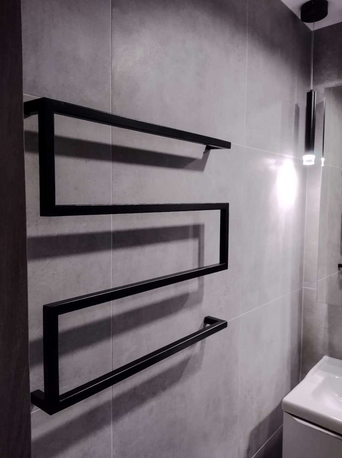 Nowoczesna łazienka z szarymi płytkami i czarnym, designerskim grzejnikiem drabinkowym, oświetlona wiszącą lampą.