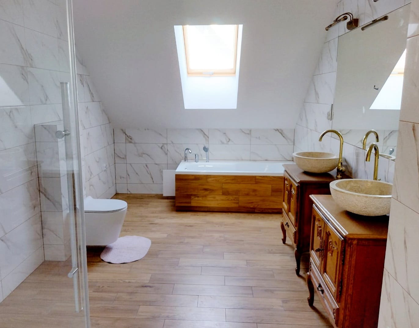 Wykończona łazienka na poddaszu z marmurowymi płytkami, drewnianą podłogą, wolnostojącą wanną z drewnianą obudową i szafkami z kamiennymi umywalkami.