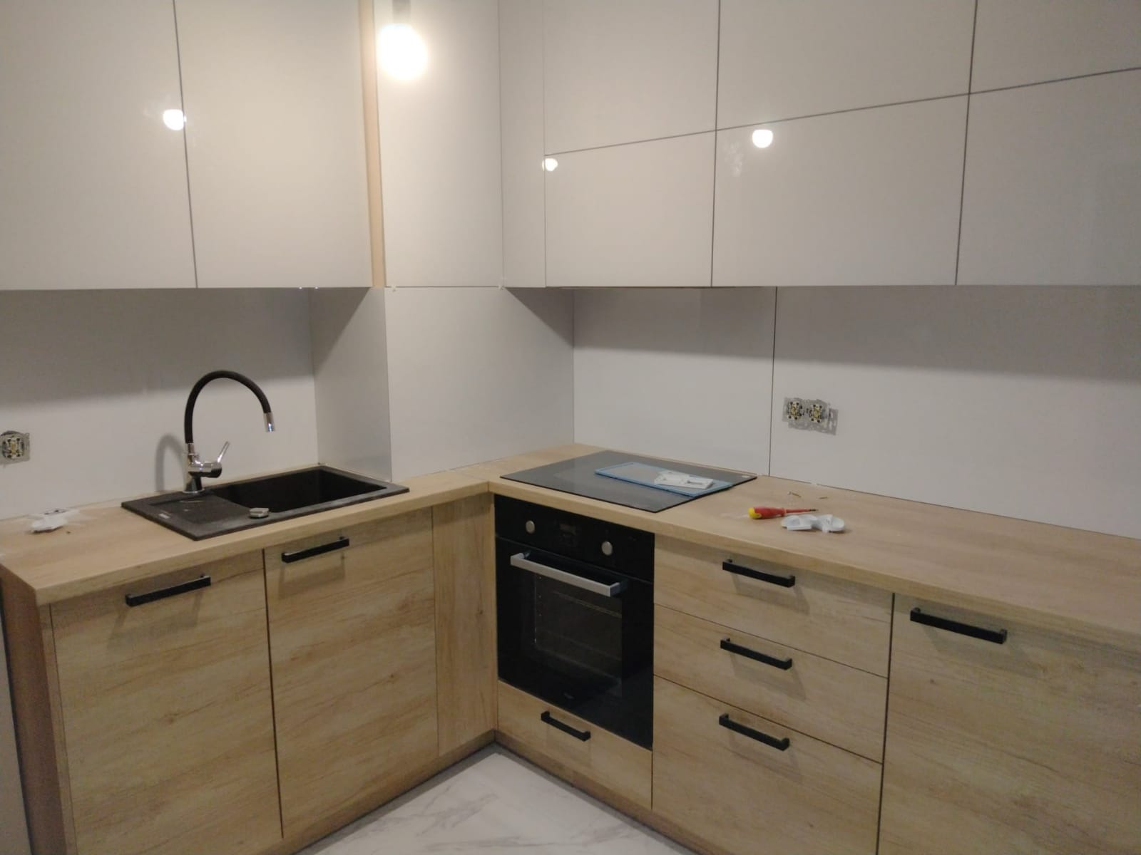 Jasna kuchnia z drewnianymi szafkami, czarnym zlewem i płytą indukcyjną, widoczne gniazdka elektryczne i narzędzia na blacie.