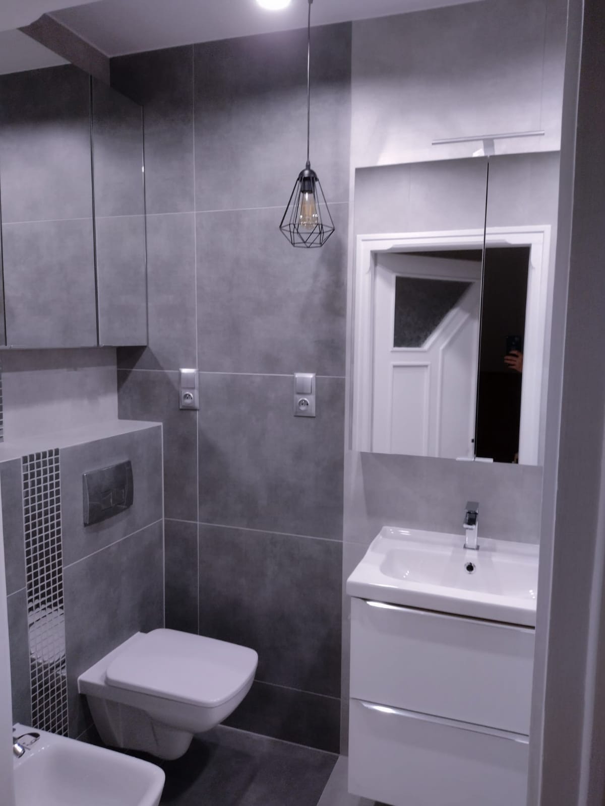 Nowoczesna łazienka w szarościach z wiszącą toaletą, umywalką z szafką i geometryczną lampą wiszącą, odbicie osoby w lustrze.