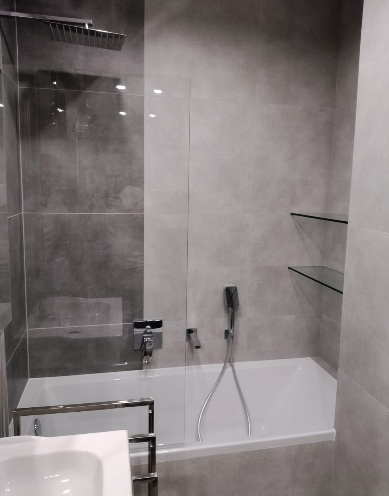 Nowoczesna łazienka wykończona w szarościach, z prostokątną kabiną prysznicową typu walk-in, chromowaną armaturą, wanną oraz szklanymi półkami na ścianie.