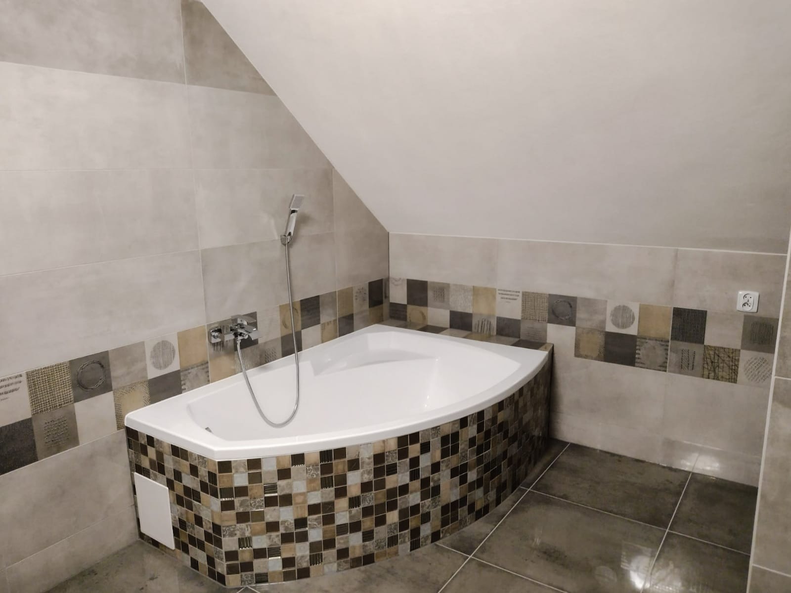 Narożna wanna obudowana mozaiką w odcieniach brązu i beżu, z chromowaną baterią i słuchawką prysznicową na tle jasnych, beżowych płytek w łazience na poddaszu.