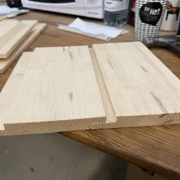 Przygotowane deski boazeryjne z wpustem i wypustem w Trzebiatowie, leżące na drewnianym stole obok młotka, butelki wody i kubka.