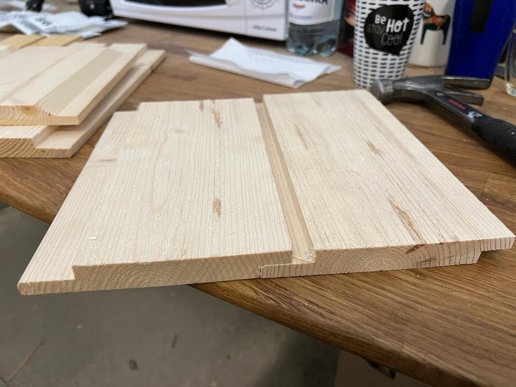 Przygotowane deski boazeryjne z wpustem i wypustem w Trzebiatowie, leżące na drewnianym stole obok młotka, butelki wody i kubka.