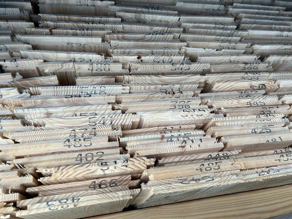 Stos ponumerowanych desek z jasnego drewna, widoczne słoje i czarne numery naniesione na końcach desek.