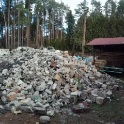 Góra gruzu ceglanego i betonowego po rozbiórce budynku, z widocznymi fragmentami zbrojenia, na tle lasu i częściowo zdemontowanego budynku gospodarczego.