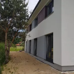 Dwukondygnacyjny dom z jasnoszarą i białą elewacją, z ciemnymi okiennymi ramami, widoczny z boku, z dużym świerkiem na pierwszym planie i piaskiem na ziemi.